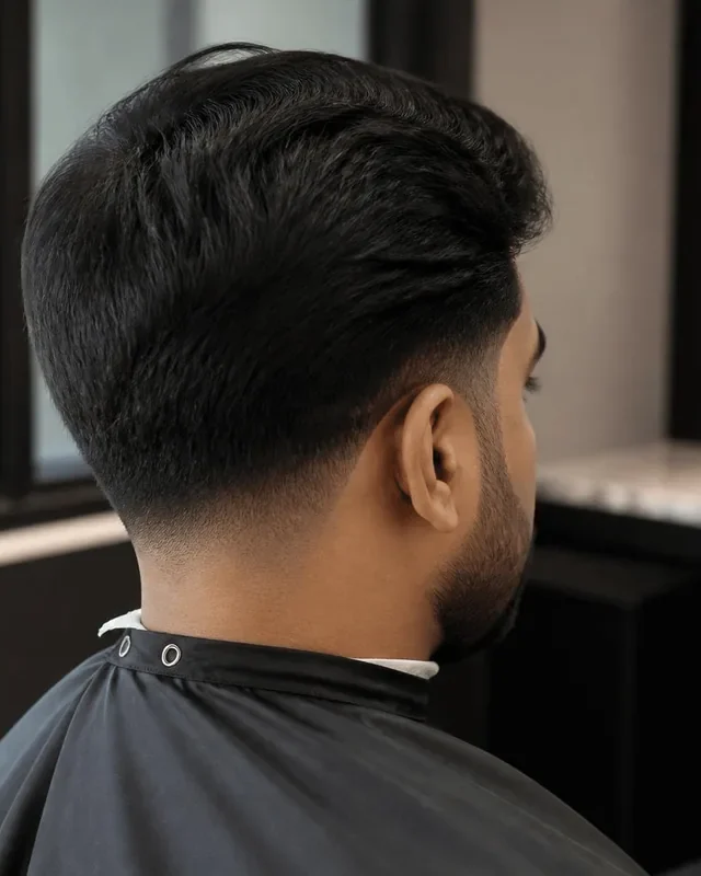 Low drop fade curving behind the ear on a South Asian man with thick black hair in a barbershop chair, soft window light